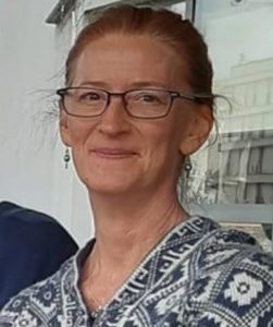Head and shoulders shot of a cis white female with dark rectangular glasses and earrings