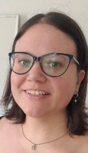 Head and shoulders shot of a smiling cis white female with round dark glasses and a necklace
