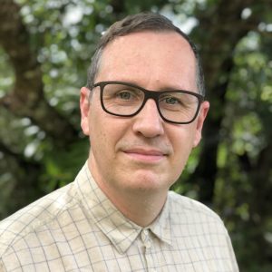 Head and shoulders shot of a cis white male with short dark hair wearing rectangular black glasses and a lightly checked short
